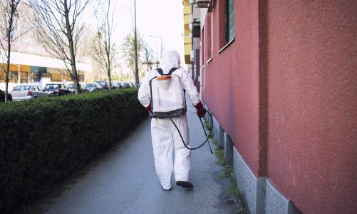 Unrecognizable worker in chemical protection suit spraying disinfectant on public surfaces.